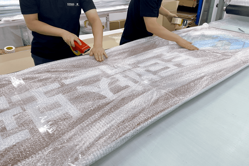 2_포장배송_11_킨코스 직원들이 대형 간판 인쇄물을 버블랩으로 감싸며 테이핑 작업을 진행하는 모습, 파손 방지를 위한 보호 포장 과정
