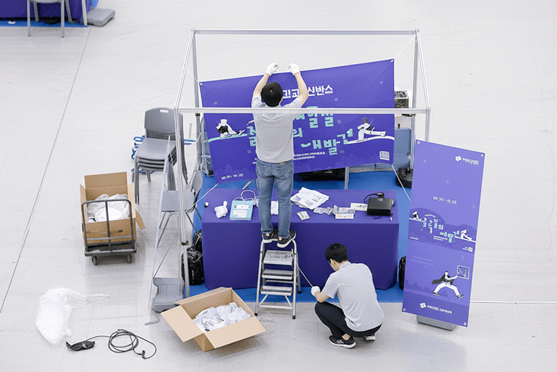 2_포장배송_17_킨코스 직원들이 전시회 부스에 배너와 POP를 설치 중인 모습, 제작물 배송 후 현장 세팅까지 진행하는 모습