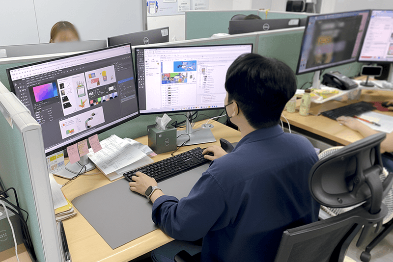 4_인력지원_15_킨코스 디자이너가 듀얼 모니터로 디자인 시안을 검토하며 고객사의 피드백을 반영한 업무 중인 모습