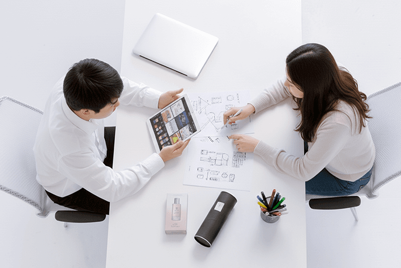 4_인력지원_7_태블릿과 종이 도면을 보며 제품 패키지 구조를 논의하는 기획자와 디자이너의 협업 장면